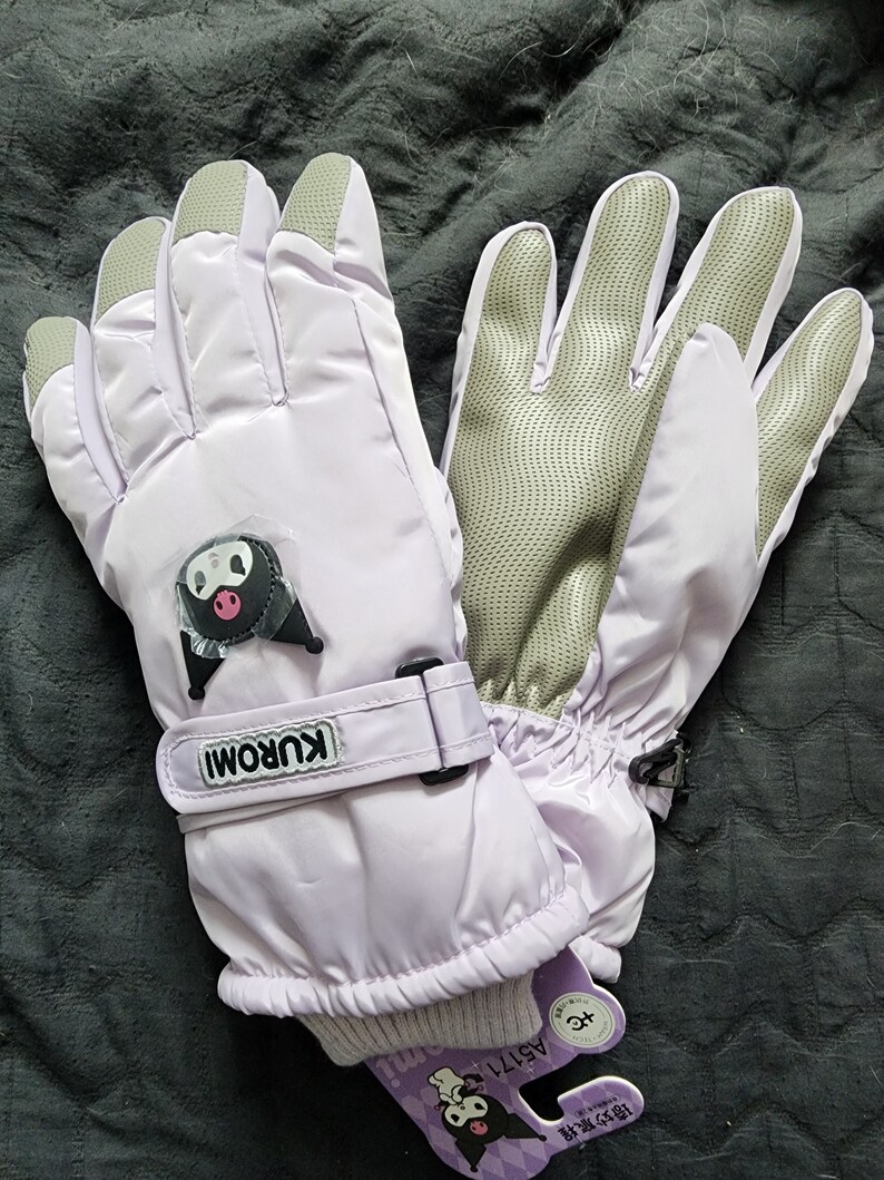 K&ouml;nnte beinhalten: Ein Paar lavendelfarbene Kinder-Winterhandschuhe mit einer schwarz-wei&szlig;en Comicfigur auf dem linken Handschuh. Die Handschuhe haben einen Klettverschluss am Handgelenk und ein schwarz-wei&szlig;es Etikett mit dem Text "KUROMI" auf dem linken Handschuh. Das Etikett tr&auml;gt auch den Text "AS1711" und eine schwarz-wei&szlig;e Comicfigur.