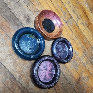 Può includere: Quattro posacenere rotondi con interni colorati e glitterati. I posacenere sono nelle tonalità del blu, del viola e del rame. I posacenere sono realizzati in resina e hanno una finitura lucida. I posacenere sono su una superficie di legno.