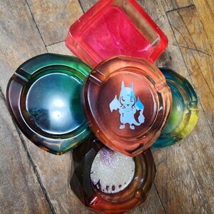 Può includere: Una collezione di posacenere colorati in resina di varie forme. I posacenere presentano una gamma di colori, tra cui rosso, verde, arancione e blu. Un posacenere ha un design di personaggi dei cartoni animati. I posacenere sono disposti su una superficie di legno.