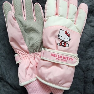 K&ouml;nnte beinhalten: Rosa Hello Kitty Winterhandschuhe mit wei&szlig;em und grauem Innenfutter. Die Handschuhe haben einen Klettverschluss mit dem Text "HELLO KITTY '23 SANRIO".