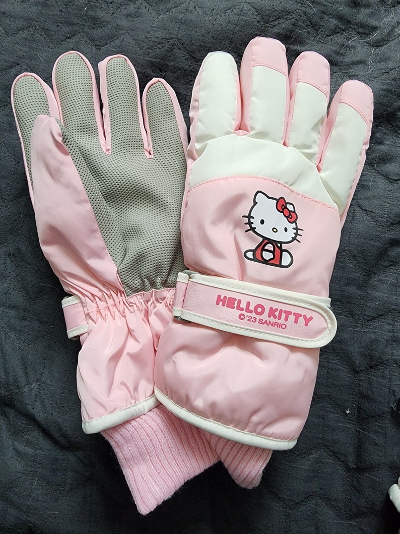 K&ouml;nnte beinhalten: Rosa Hello Kitty Winterhandschuhe mit wei&szlig;em und grauem Innenfutter. Die Handschuhe haben einen Klettverschluss mit dem Text "HELLO KITTY '23 SANRIO".