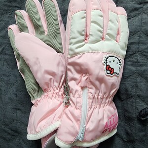 K&ouml;nnte beinhalten: Rosa Hello Kitty Skif&auml;ustlinge mit einem wei&szlig;en Rei&szlig;verschluss und einem Hello Kitty Logo auf der R&uuml;ckseite des rechten Handschuhs. Die Handschuhe haben eine graue, strukturierte Handfl&auml;che.