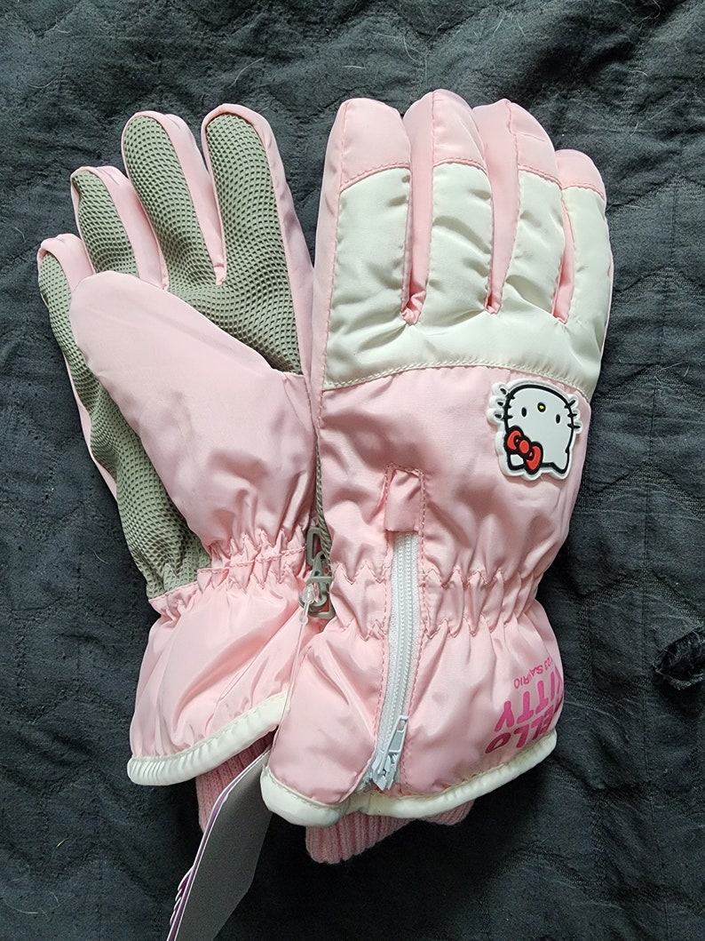K&ouml;nnte beinhalten: Rosa Hello Kitty Skif&auml;ustlinge mit einem wei&szlig;en Rei&szlig;verschluss und einem Hello Kitty Logo auf der R&uuml;ckseite des rechten Handschuhs. Die Handschuhe haben eine graue, strukturierte Handfl&auml;che.