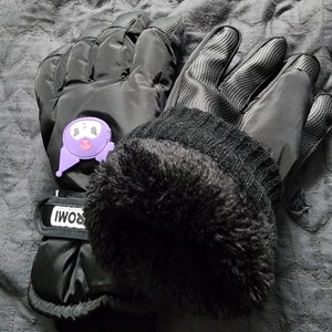 K&ouml;nnte beinhalten: Ein Paar schwarze Winterhandschuhe mit einem lilafarbenen Comic-Gesicht auf einem Handschuh. Die Handschuhe haben ein Fleecefutter und einen schwarzen Riemen mit dem Wort "ROMI" darauf.