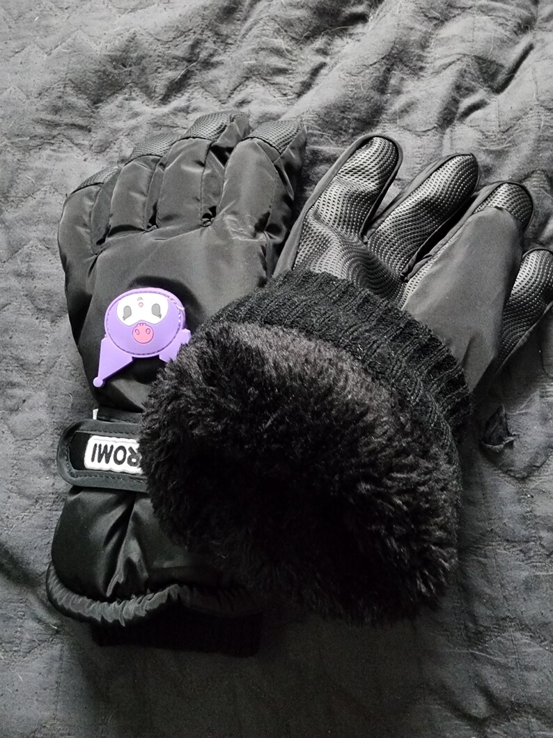 K&ouml;nnte beinhalten: Ein Paar schwarze Winterhandschuhe mit einem lilafarbenen Comic-Gesicht auf einem Handschuh. Die Handschuhe haben ein Fleecefutter und einen schwarzen Riemen mit dem Wort "ROMI" darauf.