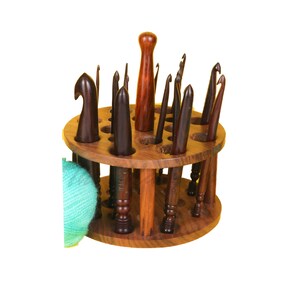 Op de afbeelding: Een houten haaknaaldenhouder met een ronde basis en meerdere sleuven om haaknaalden op te bergen. De houder is gevuld met haaknaalden in verschillende maten. Een bol teal-kleurig garen bevindt zich op de voorgrond.