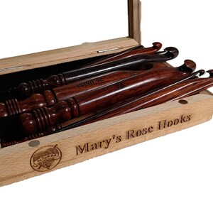 Op de afbeelding: Een set houten haaknaalden in een houten doos met een glazen deksel. De doos is gegraveerd met "Mary's Rose Hooks".