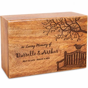 Op de afbeelding: Een rechthoekige houten urn met een lasergegraveerd ontwerp van een parkbank onder een boom met een vogel op de rugleuning. De tekst luidt: in Loving Memory of Darrelle E. Arthur JULY 10, 1976 - AUGUST 4, 2023.