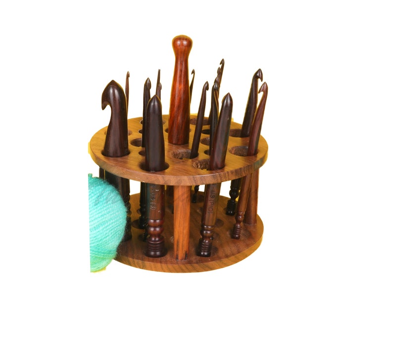 Op de afbeelding: Een houten haaknaaldenhouder met een ronde basis en meerdere sleuven om haaknaalden op te bergen. De houder is gevuld met haaknaalden in verschillende maten. Een bol teal-kleurig garen bevindt zich op de voorgrond.