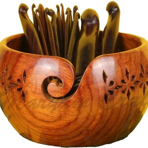 Op de afbeelding: Een houten garenkom met een gesneden bloemenpatroon en een ingebouwde garengeleider. De kom is gevuld met een set houten haaknaalden.
