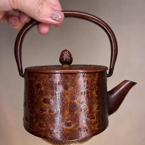 May include: A brown, textured teapot with a cylindrical body, a curved handle, and a small spout. The lid features a decorative knob, and the exterior surface has a mottled, rustic finish with a darker, hammered-style texture on the base.