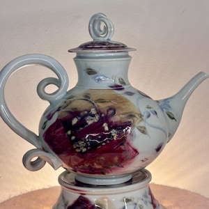 May include: A white ceramic teapot with a matching base, featuring a hand-painted design of deep red and purple floral motifs. The teapot has a curved handle with a scroll detail, a tapered spout, and a lid topped with a decorative swirl handle.