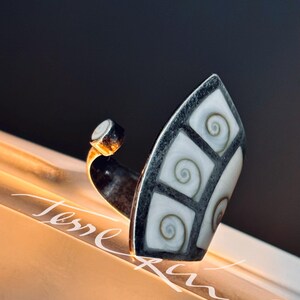 May include: A silver-toned ring featuring a large, shield-shaped face inlaid with three white, square-cut shell segments, each displaying a natural brown spiral pattern. The metal band has a dark, oxidised finish and a small, circular shell accent.