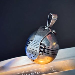 May include: A spherical silver pendant featuring a hinged mechanism with horizontal grooved lines and a small perforated plate detail. The metal surface has a polished finish with a metallic sheen, and a sturdy bail is attached to the top.