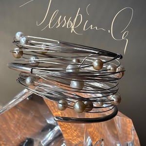 May include: A silver-toned wire bracelet with multiple strands, adorned with cream and light brown pearls. The bracelet is wrapped around a clear, faceted object. The image includes the handwritten text "Tessa Kim".
