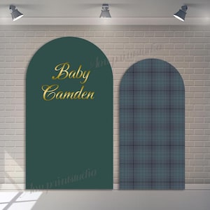 May include: Two arched backdrop panels. One is dark teal with gold script reading "Baby Camden." The other panel has a blue and grey plaid pattern. The backdrop is set against a white brick wall, illuminated by overhead spotlights.
