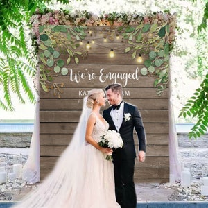 Peut inclure: Une toile de fond en bois rustique avec de la verdure et des guirlandes lumineuses, décorée du texte "We're Engaged". Un couple se tient devant la toile de fond, la mariée portant une robe de mariée blanche et le marié portant un costume noir.