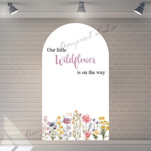 May include: A white arched backdrop with the text "Our little Wildflower is on the way" in pink script. The bottom of the backdrop features a watercolor floral design with pink, yellow, blue, and purple flowers. The backdrop is set against a brick wall.