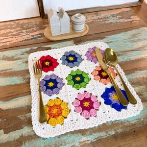 Puede incluir: Un mantel individual cuadrado de ganchillo blanco con nueve motivos florales coloridos. Cada flor tiene un color diferente, incluyendo rojo, azul y amarillo. Un tenedor, un cuchillo y una cuchara dorados están colocados sobre el mantel individual.