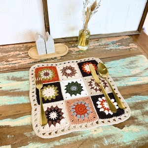 Può includere: Un sottopentola quadrato all'uncinetto multicolore con un motivo floreale. Il sottopentola presenta quadrati marroni, arancioni, verdi e bianchi, con un bordo beige. Una forchetta, un coltello e un cucchiaio dorati sono posizionati sul sottopentola.