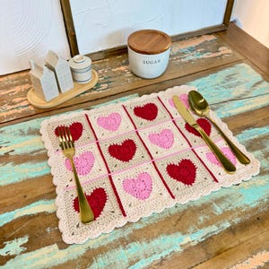 Puede incluir: Un mantel individual de ganchillo con un patrón de corazones rojos y rosas sobre un fondo beige. El mantel individual tiene un borde festoneado y se muestra con cubiertos dorados. Un azucarero y saleros y pimenteros están en el fondo.
