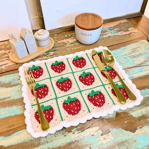 Puede incluir: Un mantel individual de ganchillo con un patrón de fresas rojas con hojas verdes sobre un fondo crema. Un tenedor, un cuchillo y una cuchara dorados están colocados encima. El diseño está enmarcado por un borde blanco festoneado.