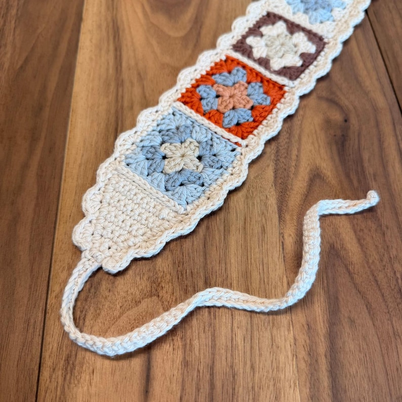 Close-up of a rustic granny square crochet headband showing blue, rust, and neutral floral motifs with scalloped edging and tie-back strings on a wooden surface.