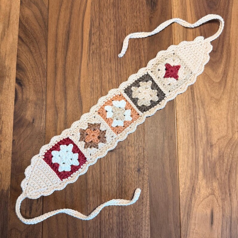 Rustic granny square crochet headband laid flat on a wooden surface, featuring connected floral motifs in neutral and earthy tones with scalloped edging and tie-back ends.