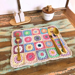 May include: A crocheted placemat with a colorful floral pattern. The placemat is made of squares with different colored flowers in each square. The placemat is on a wooden table with a fork and knife on top.
