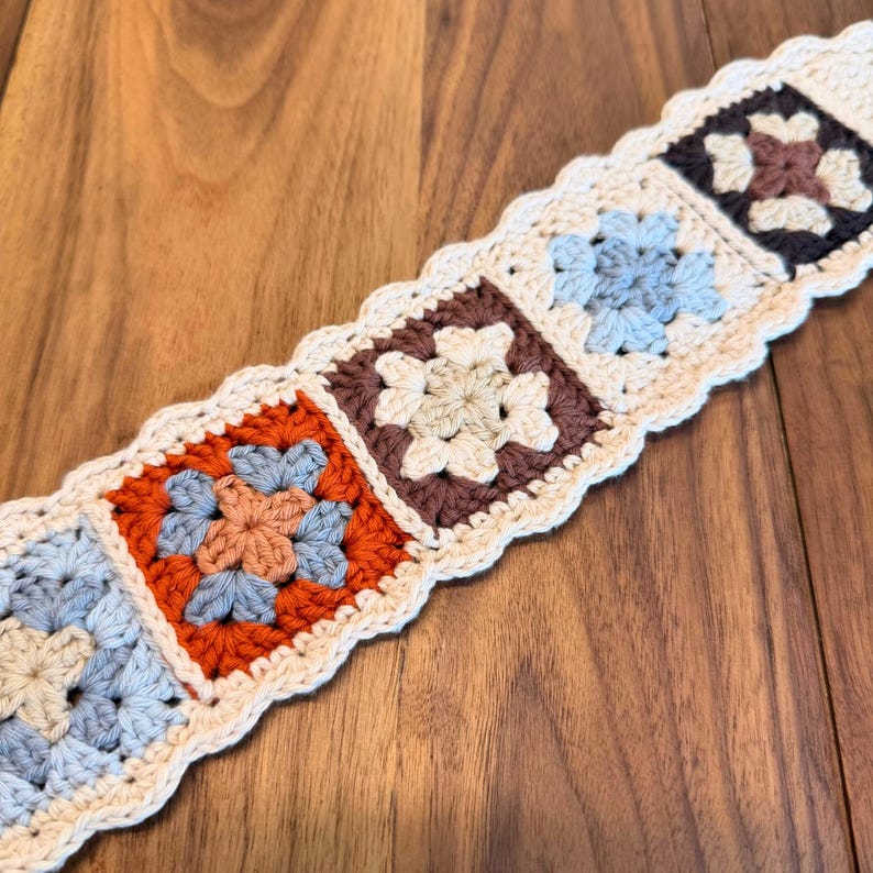 Close-up of connected floral granny squares in a rustic crochet headband, showing repeating motifs in blue, rust, brown, and cream yarn on a wooden surface.