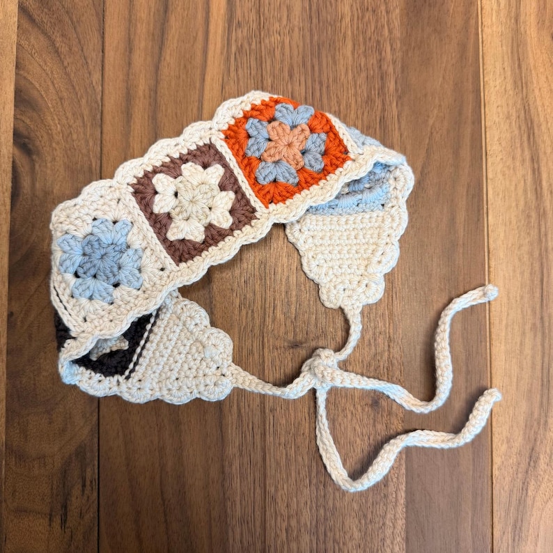 Rustic granny square crochet headband partially folded on a wooden surface, showing floral motifs, scalloped edging, and adjustable tie-back strings.