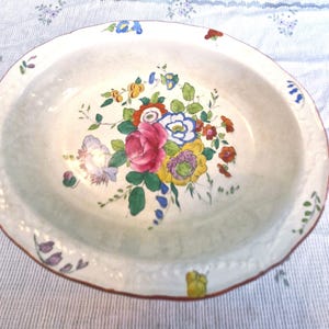 Könnte beinhalten: Ovale, cremefarbene Servierschale aus Keramik mit floralem Design. Die Mitte zeigt einen Strauß aus rosa, roten, gelben, blauen und weißen Blumen mit grünen Blättern. Der Rand hat einen gewellten Rand und einen braunen Rand.