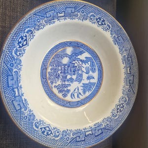 Könnte beinhalten: Zwei weiß-blaue Keramikplatten mit Goldrand. Die Teller weisen ein detailliertes blau-weißes Willow-Muster auf. Die Teller sind rund und haben eine leicht tiefe Schüsselform.