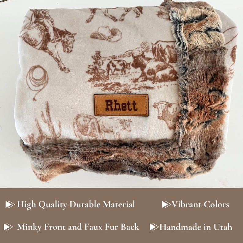 Puede incluir: Una manta para beb&eacute; de color crema con un estampado marr&oacute;n con tem&aacute;tica de vaqueros, que incluye vaqueros, caballos y ganado. La manta tiene un ribete de piel sint&eacute;tica y un parche de cuero que dice "Rhett".