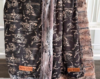 May include: Two plush blankets with a black background and a cowboy-themed design. The blankets have a faux fur trim in shades of brown and gray. One blanket has a leather tag that reads "Murray LaVerne" and the other reads "Beau William".