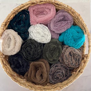 May include: A woven basket filled with various colors of faux fur yarn. The yarn is in shades of teal, pink, purple, white, black, green, brown, and tan. The basket is made of natural woven material.