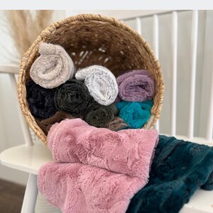 May include: A woven basket overflowing with rolled plush blankets in various colors, including pink, teal, black, and gray. The blankets are soft and textured, with some spilling out of the basket onto a white surface.
