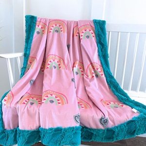 May include: A pink blanket with a rainbow pattern and teal faux fur trim. The blanket features a repeating design of colorful rainbows and hearts. The blanket is draped over a white chair.