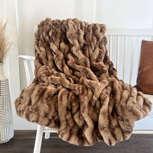 May include: A brown faux fur throw blanket draped over a white chair. The blanket has a textured, wavy pattern and a warm, neutral colour. A decorative vase with tall, dried grass and a brown leather cushion are also visible.