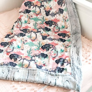 May include: A soft, plush baby blanket with a gray faux fur border. The blanket features a pink, green, and white pattern with black silhouettes of buffalo, cacti, and flowers. The words "Wild" are repeated throughout the pattern.
