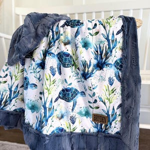May include: A baby blanket featuring a sea turtle pattern in shades of blue, green, and white. The blanket has a plush, navy blue border and a matching hood. A brown leather tag displays the name "BRYDEN".