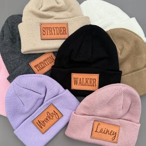 May include: A collection of beanies in various colors, including beige, black, white, and pink. Each beanie has a rectangular leather patch with a name engraved on it. The beanies are made of a soft, knitted material.