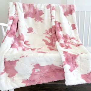 May include: A soft, plush blanket in white with pink tie-dye-style patterns. The blanket is draped over a white chair, showcasing its texture and design. The cozy throw blanket is perfect for adding warmth and style to any space.