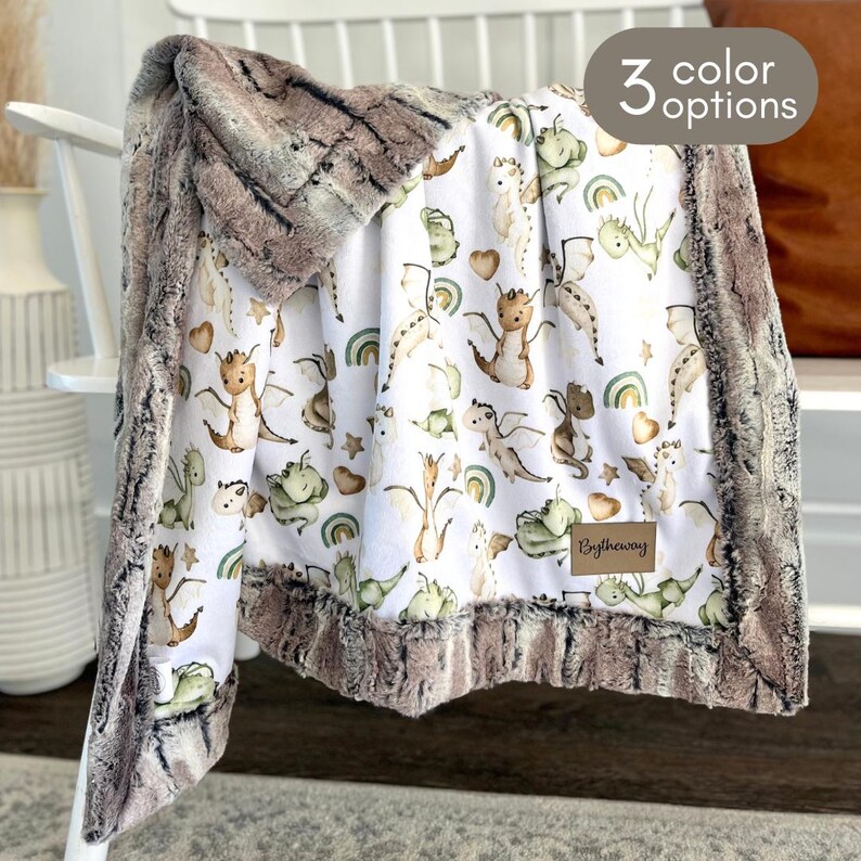 Puede incluir: Una manta de beb&eacute; de felpa con un fondo blanco con un estampado de dragones amigables, arco&iacute;ris y corazones en tonos verdes, marrones y beige. La manta tiene un ribete de piel sint&eacute;tica marr&oacute;n y el texto "3 color options". Una peque&ntilde;a etiqueta dice "Bytheway".