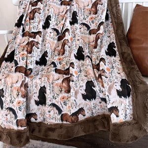 May include: A soft, white blanket featuring a repeating pattern of horses in various shades of brown and black, interspersed with orange flowers and green foliage. The blanket has a brown, plush border, creating a cozy and inviting aesthetic.