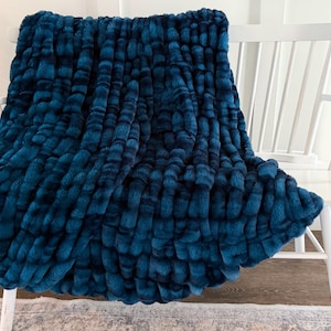 May include: A deep teal blue faux fur throw blanket with a textured, grid-like pattern of raised, plush squares. The blanket features a thick, soft pile and a consistent, rich color throughout, draped over a white wooden bench.
