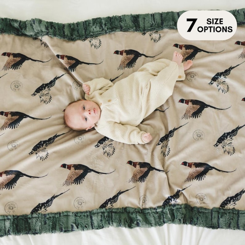 May include: A baby lying on a beige and green plush blanket with a pheasant print. The blanket has a green faux fur trim. The text "7 SIZE OPTIONS" is in the upper right corner of the image.