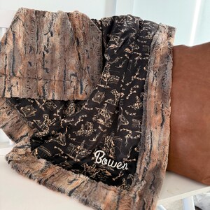 Puede incluir: Una manta suave en marr&oacute;n y negro con un dise&ntilde;o de vaquero y el nombre "Bowen" bordado en blanco. La manta tiene un ribete de piel sint&eacute;tica y est&aacute; sobre una superficie blanca.