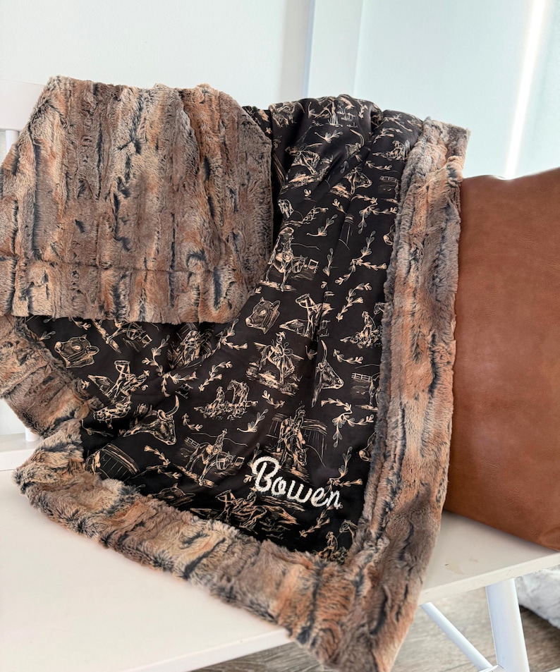 Puede incluir: Una manta suave en marr&oacute;n y negro con un dise&ntilde;o de vaquero y el nombre "Bowen" bordado en blanco. La manta tiene un ribete de piel sint&eacute;tica y est&aacute; sobre una superficie blanca.