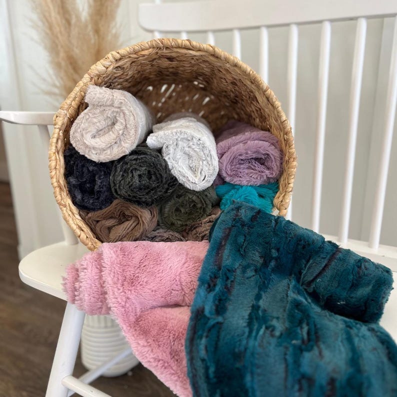 May include: A woven basket overflowing with rolled plush blankets in various colors, including pink, teal, gray, and purple. The blankets are arranged on a white chair, with additional blankets draped over the side. The textures and colors create a cozy, inviting display.